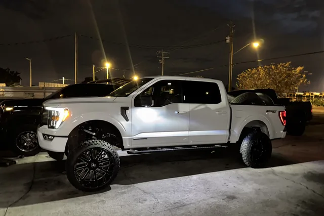 A white Ford F-150 on a lift kit and black wheels, photographed at the shop at night