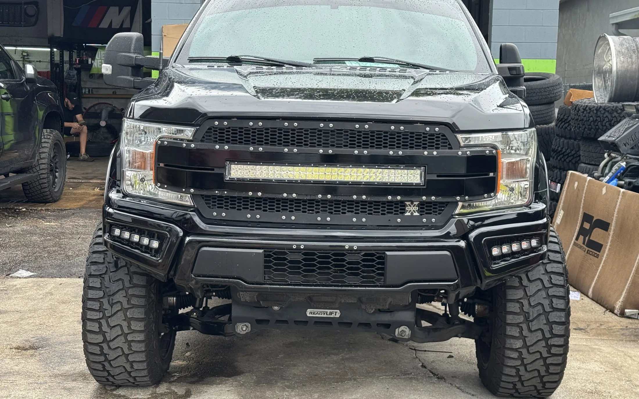 A black lifted Ford pickup with a light bar in the grille, nose on, in front of the open shop bay