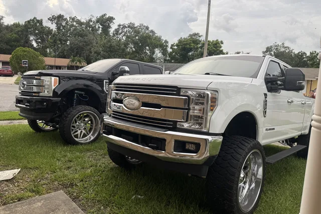 A white Ford Super Duty on chrome forged wheels, parked on the grass beside a black one
