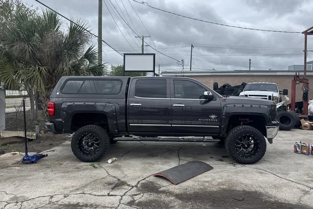 A grey Chevy Silverado with a bed cap, lifted on black wheels, side on at the shop