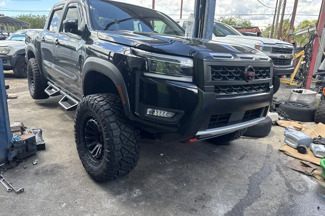 A black Nissan crew cab pickup lifted on black off-road wheels and mud-terrain tires in the shop