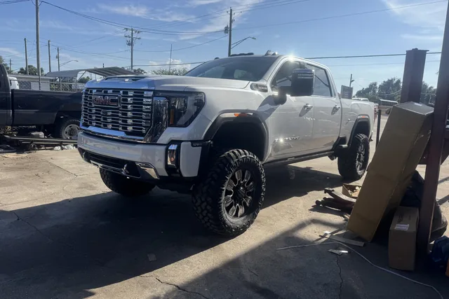 A white GMC Sierra Denali HD on a lift kit and black wheels in front of the shop