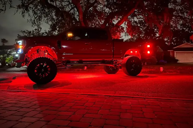 A lifted Ford Super Duty at night with red rock lights flooding the driveway under it