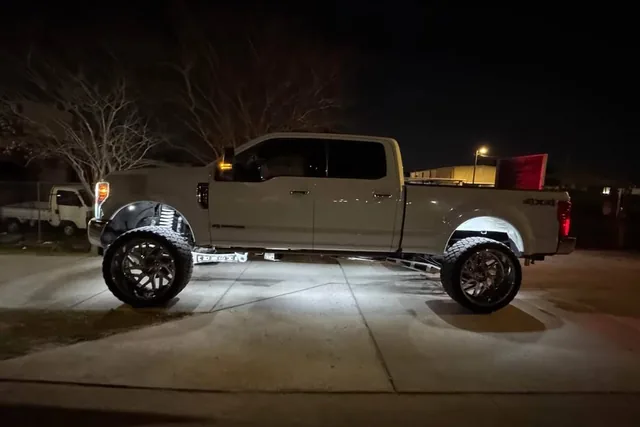 A tan Ford Super Duty at night with rock lights lighting the ground under it, side on